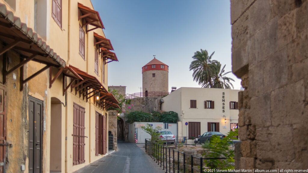 Narrow old-town street leading toward a cylindrical stone tower with a red roof and nearby palm.
