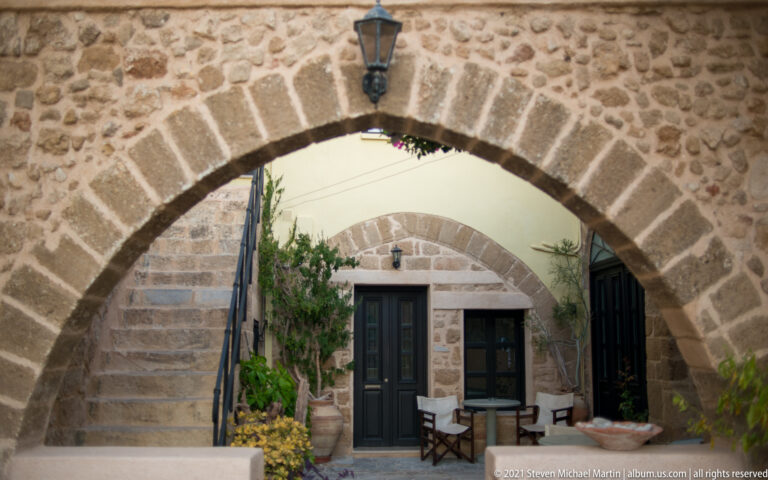 Broad stone arch framing stairs, dark doors, seating, and potted plants in a small courtyard.