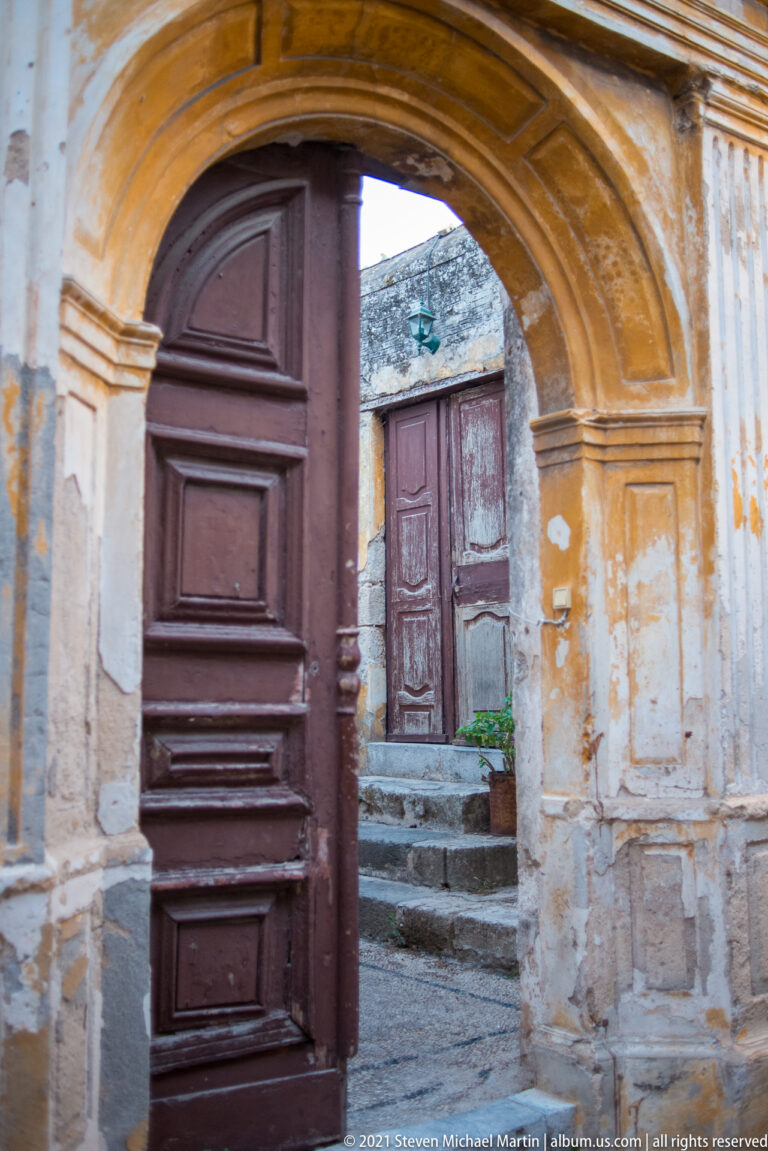 Open dark wooden door beneath a weathered molded arch, with steps leading to another door beyond.