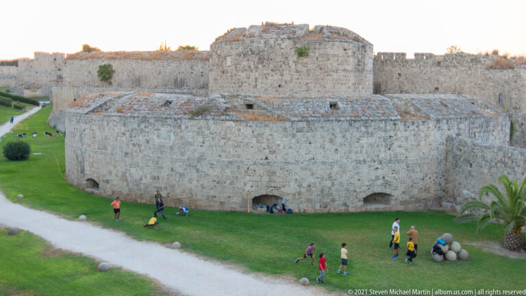 Rounded stone bastion and adjoining fortified walls above a grassy area with several visitors.