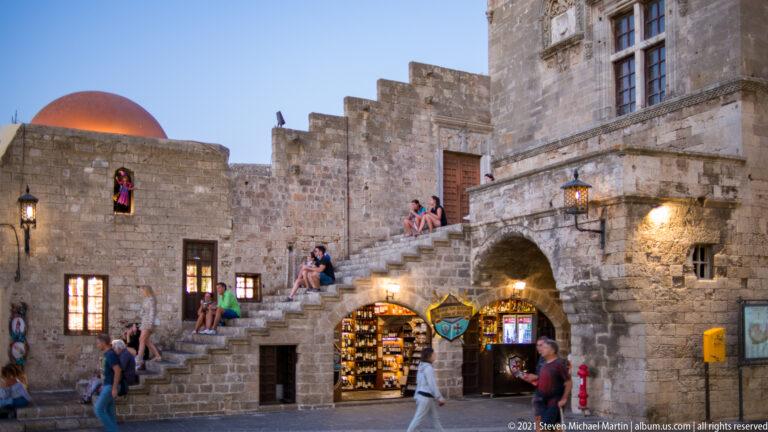 Stone complex with exterior stairs, crenellated walls, a red dome, visitors, and lit shops at twilight.