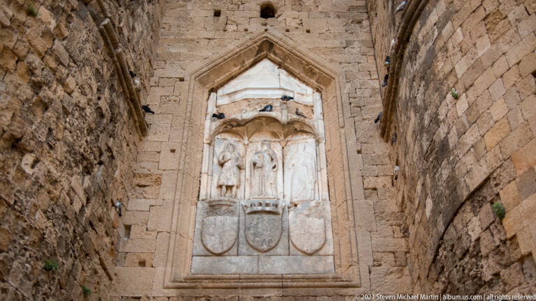 Pointed stone niche containing standing carved figures above several heraldic shields in a fortified wall.