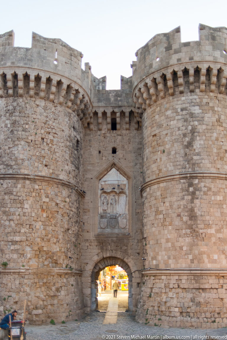 Twin round stone towers with corbelled battlements flank an arched gateway beneath a large heraldic relief.