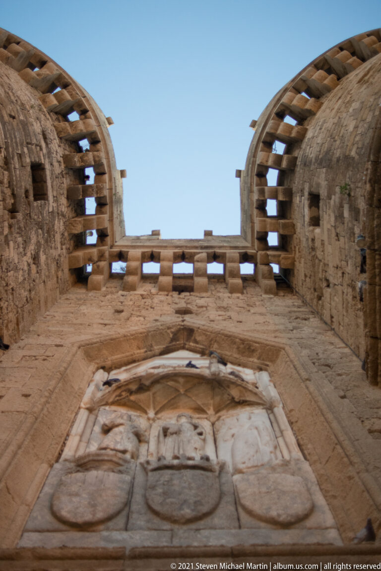 Low-angle view of heraldic shields and carved figures beneath the upper edge of a stone fortification.