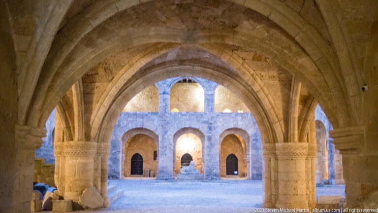Pointed stone arches and ribbed vaulting framing a two-level arcaded courtyard or hall.