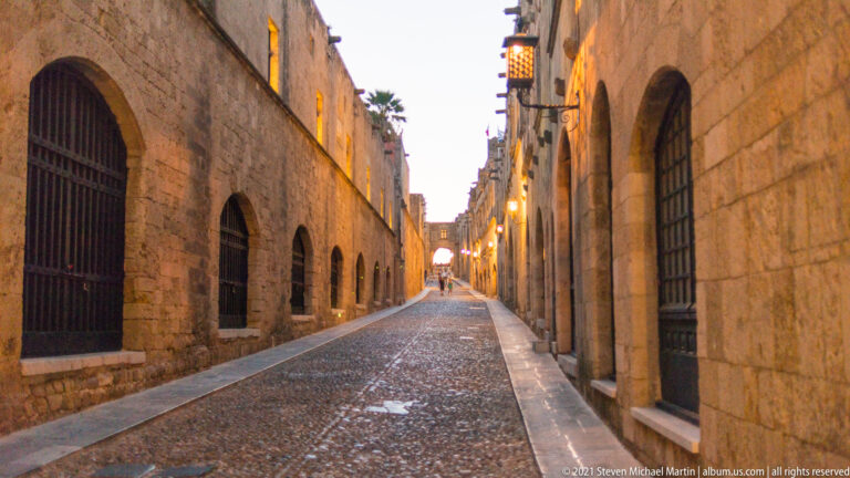 Long narrow cobbled lane between tall stone façades with arched openings and glowing lanterns at dusk.