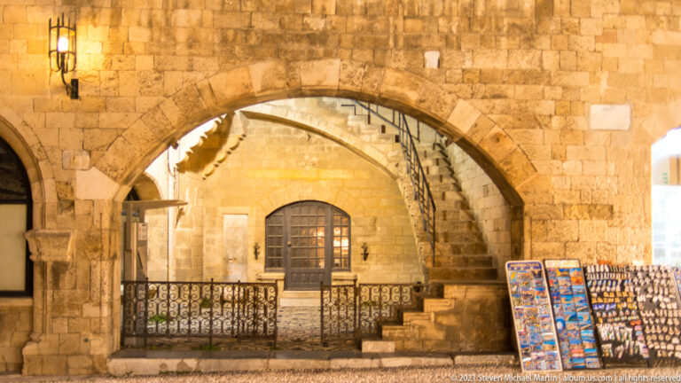 Broad pointed stone arch framing an exterior staircase, railing, and barred window, with souvenir displays nearby.