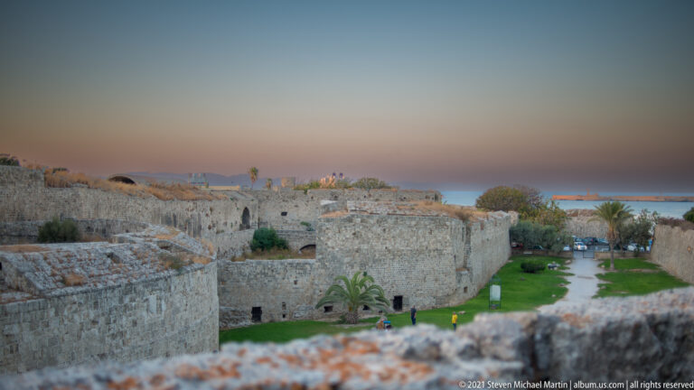 Layered stone fortifications, grassy enclosures, palms, and a path seen from above at dusk.