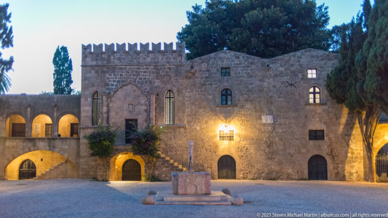 Evening courtyard bordered by a crenellated stone building with arches, stairs, and a central stone feature.