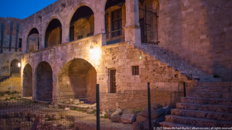 Stone staircase and arches beside a fenced area of exposed masonry remains under evening light.