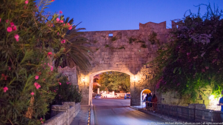Large illuminated arched gateway through a stone wall, with roadway, palms, flowering vegetation, and pedestrians.
