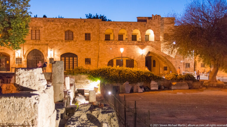 Low archaeological stone remains in front of an illuminated medieval façade and open courtyard at dusk.
