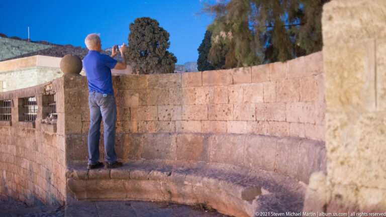 Man standing on a low stone ledge photographing over a curved masonry parapet.