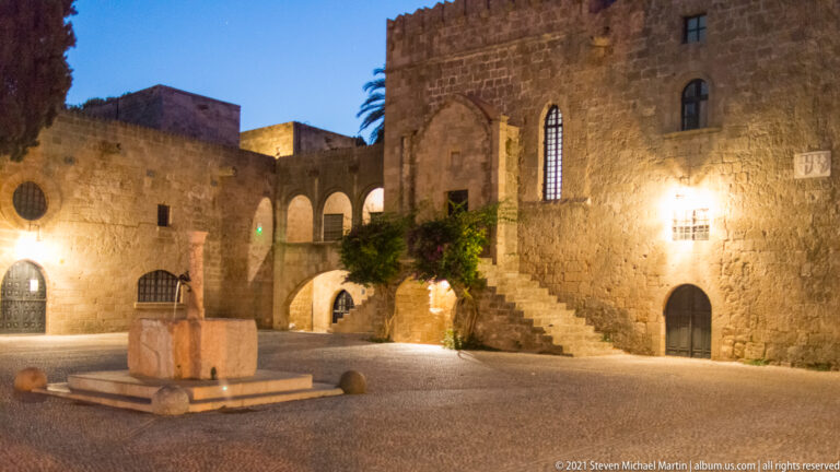 Open courtyard with a central stone feature, crenellated building, pointed window, stairs, and glowing arches.