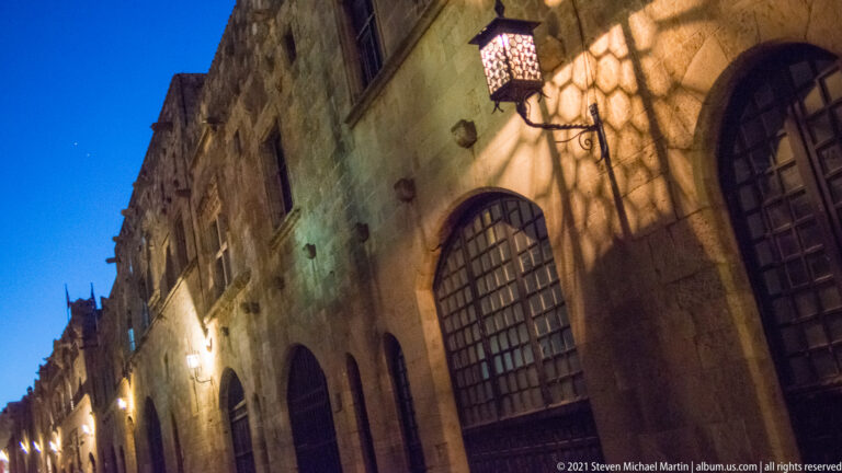 Decorative lantern casting patterned shadows across a stone façade with tall grilled arched windows.
