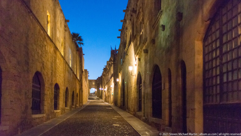 Long empty stone lane at blue hour with arched windows, metal grilles, and glowing wall lamps.