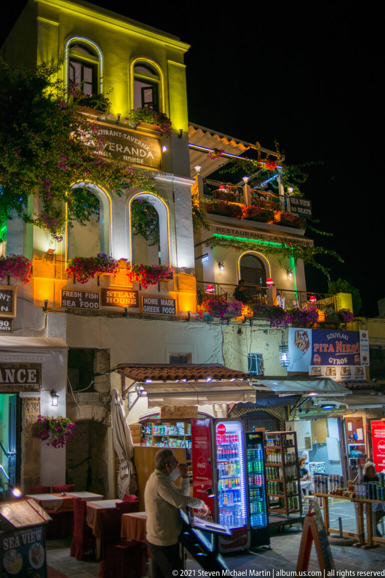 Bright souvenir and convenience storefront at night beneath arched upper windows and flower-filled balconies.