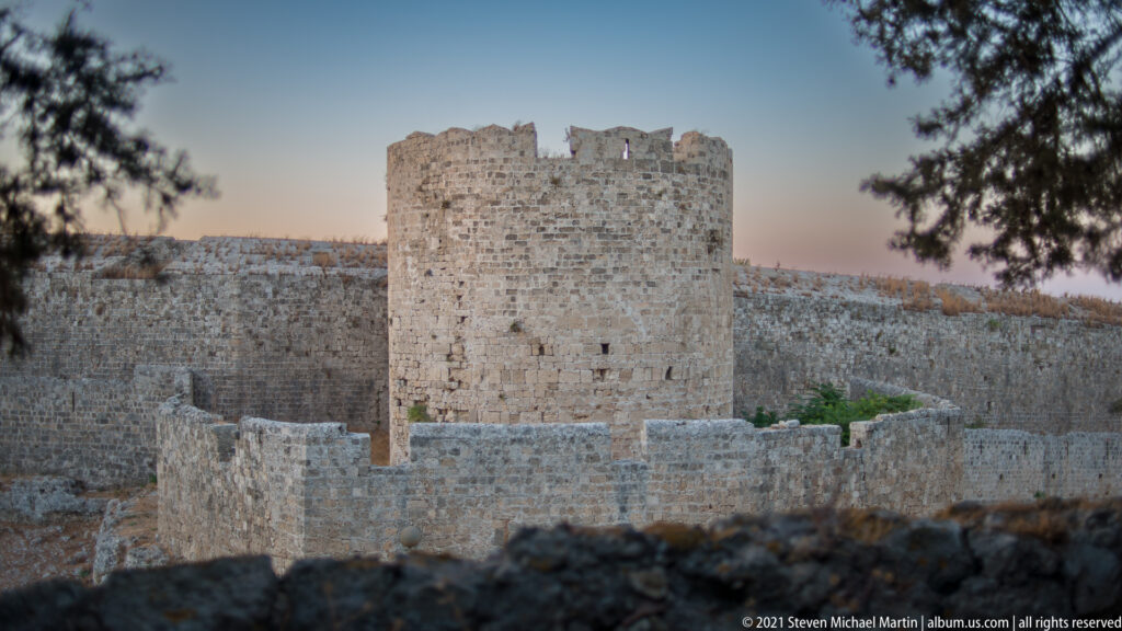 Square stone defensive tower rising above lower curtain walls, partly framed by trees.