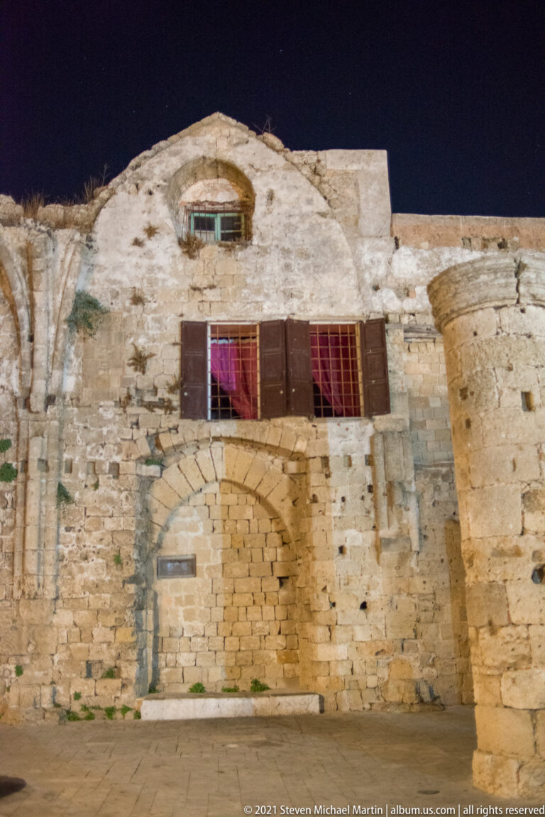 Weathered stone façade at night with a large arched opening, shuttered upper windows, and irregular masonry.