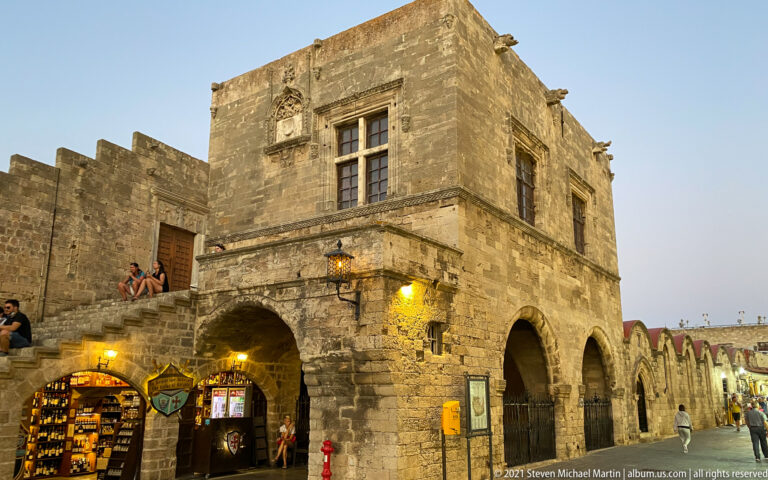 Square stone building with a large upper window, arched ground-floor passages, shops, and pedestrians at dusk.