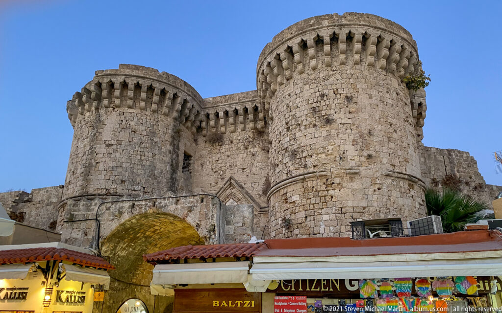 Twin cylindrical stone towers with crenellated tops flanking a narrow fortified gateway above modern shops.