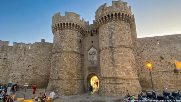 Frontal view of a fortified gateway between two cylindrical crenellated stone towers with visitors below.