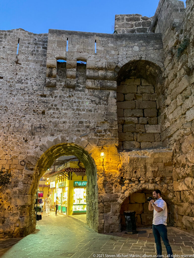 Low arched passage through a massive stone wall with bright shops beyond and blocked openings beside it.