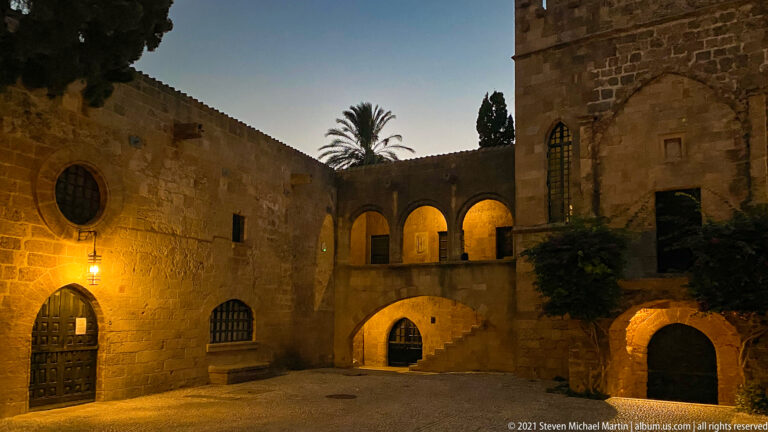 Quiet stone courtyard at night with a round window, arched doors, upper gallery, and exterior staircase.