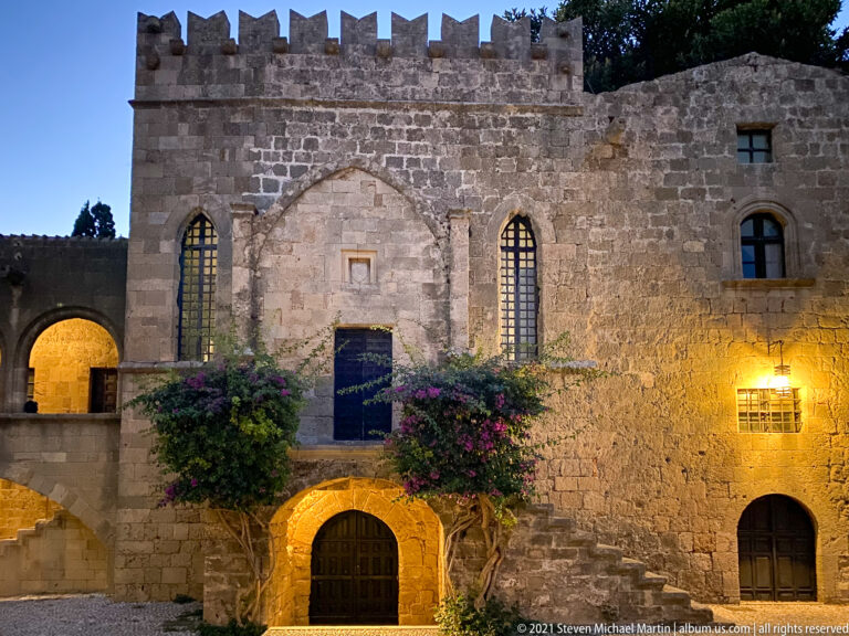 Stone façade with a pointed central recess, tall narrow windows, flowering vines, and an arched doorway.