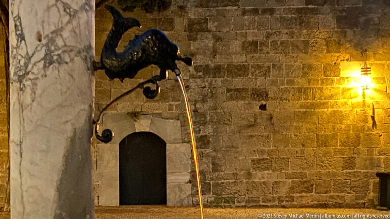 Dark animal-head fountain spout projecting from pale stone and pouring a thin stream of water.