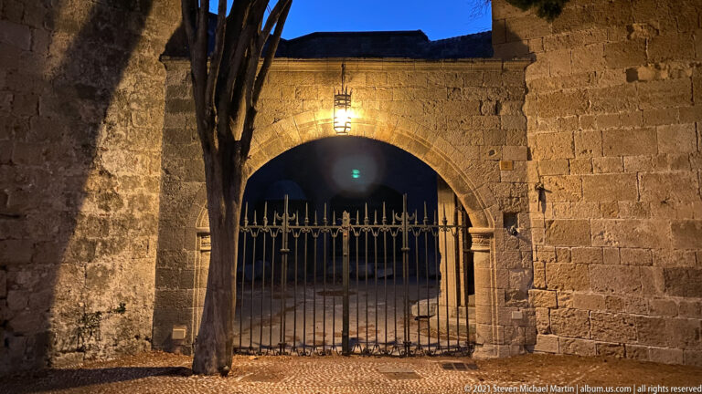 Large stone arch closed by a wrought-iron gate, lit by a hanging lantern at night.