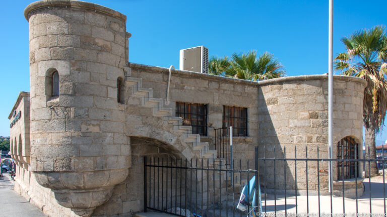 Compact stone building with a rounded corner tower, exterior stair, iron fence, and palm trees behind.