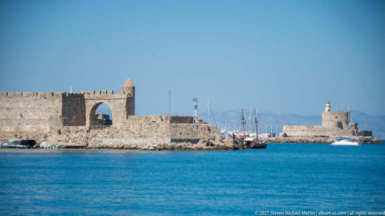 Stone harbor fortifications with a large arch, small turret, marina masts, and blue water in front.