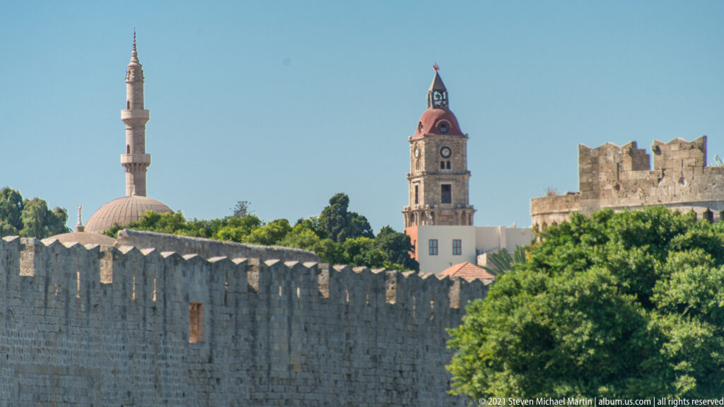 Mosque minaret and dome, red-roofed clock tower, trees, and crenellated stone walls beneath a clear blue sky.