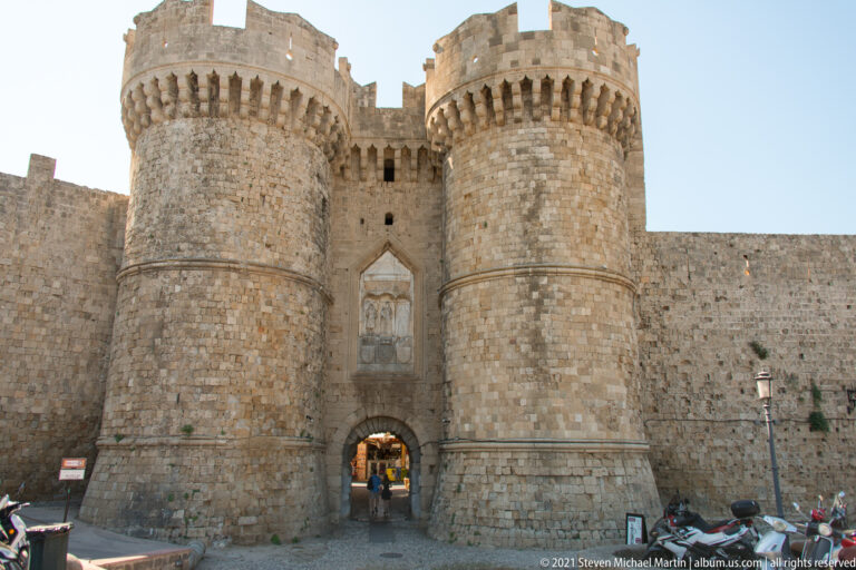 Monumental fortified gate between two massive round stone towers with corbelled battlements.