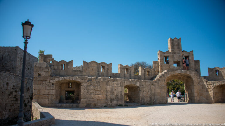 Roofless crenellated stone structure with multiple openings and a central arched passage under bright sun.