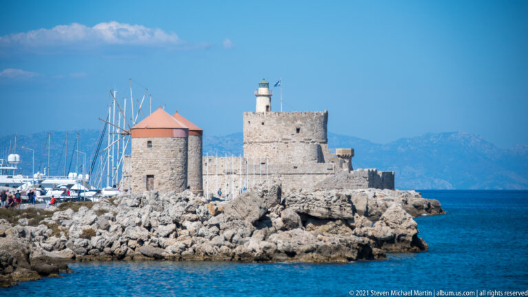 Stone windmill with red conical roof beside a squat lighthouse complex on the rocky harbor edge.