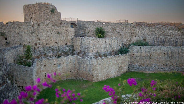 Magenta flowers in front of layered crenellated stone walls enclosing a grassy area.