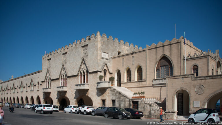 Long stone civic building with arcades, pointed windows, exterior stairs, crenellations, and parked cars.