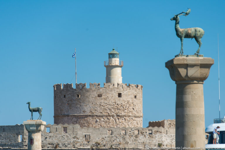 Bronze deer statue atop a stone column with a crenellated harbor lighthouse and second deer column behind.