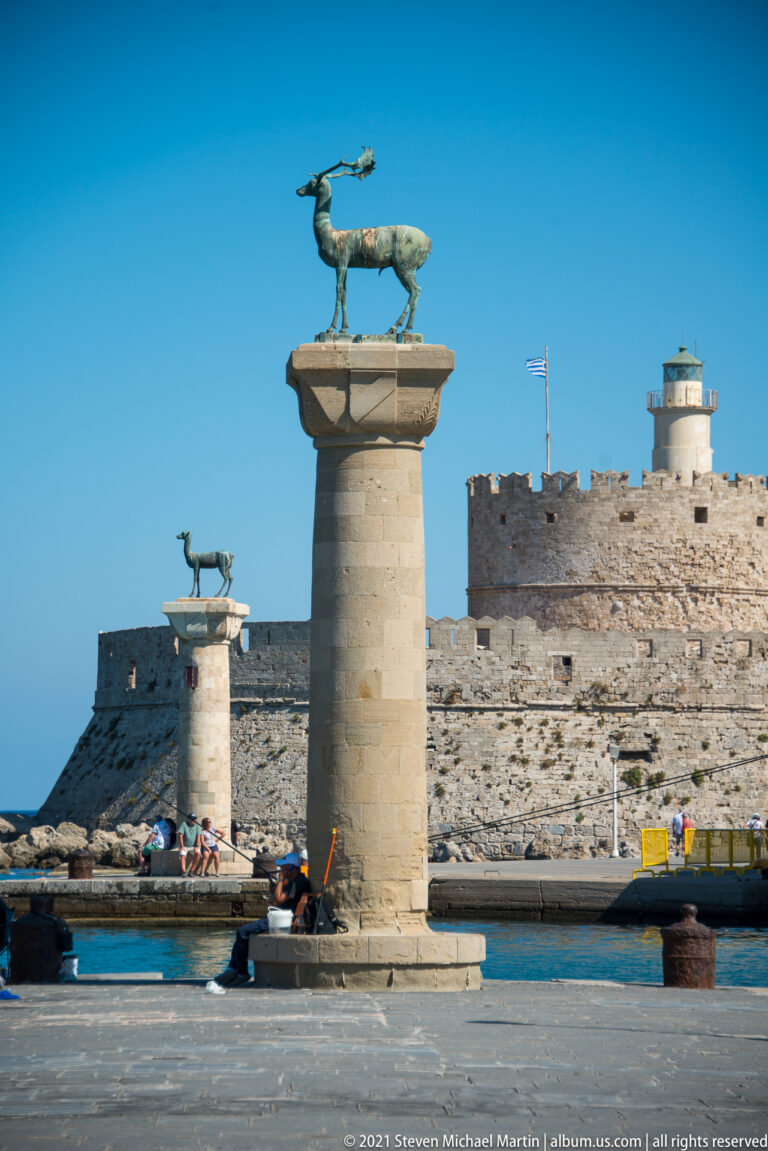 Two bronze deer statues atop stone columns beside the harbor, with a crenellated lighthouse beyond.