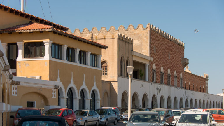Long ochre-and-white waterfront building with ground-floor arcades, pointed windows, balconies, and parked cars.