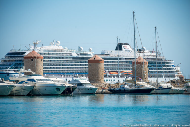 Stone windmills with red roofs among yachts and sailboats, with a large cruise ship behind.