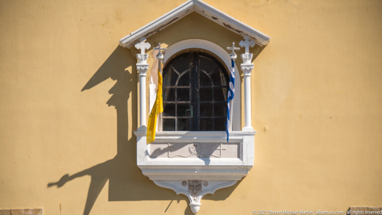 Arched dark window in an ornate white surround on an ochre wall, with two narrow flags.