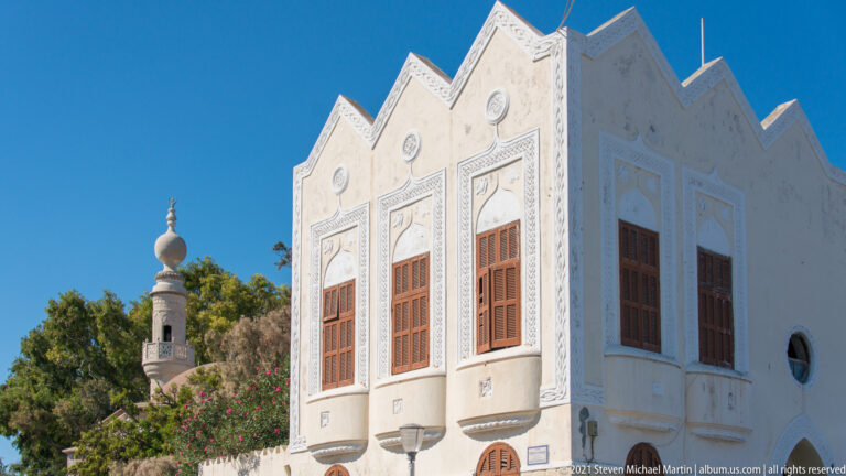 White ornate building with stepped gables, tall wooden windows, carved panels, and a slender tower behind.