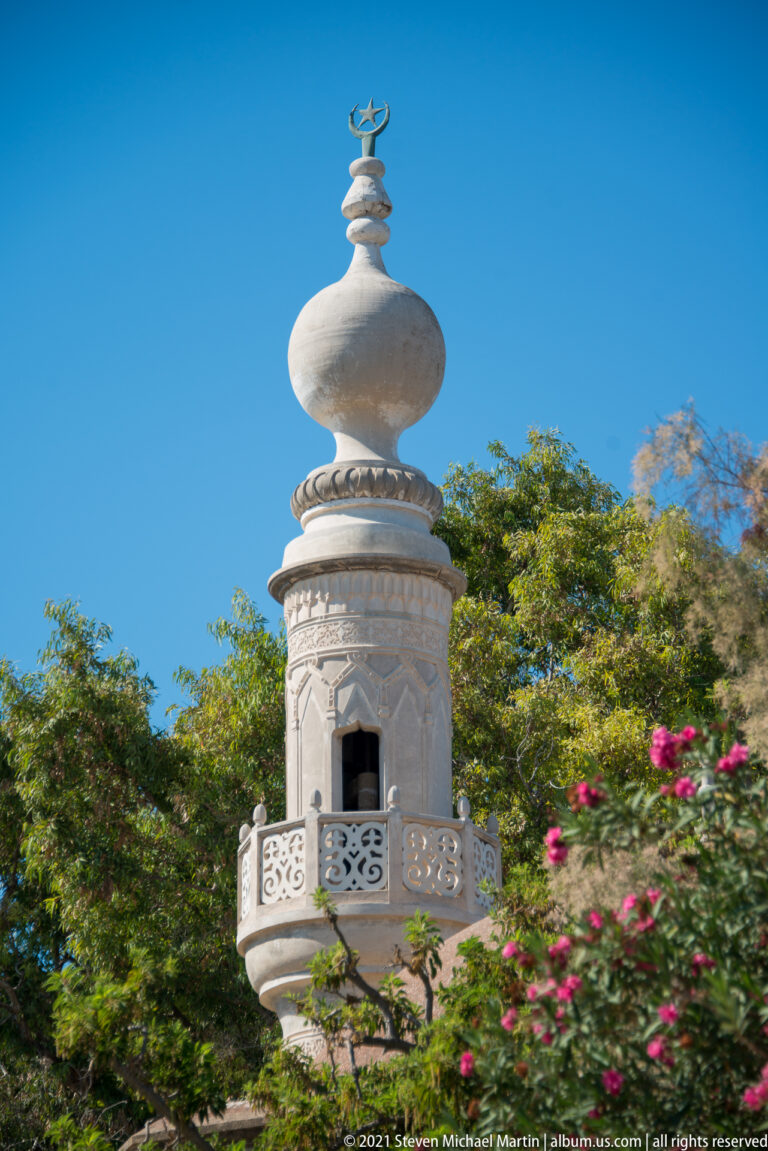 Small white tower with a perforated balcony, bulbous dome, and crescent finial above trees and flowers.