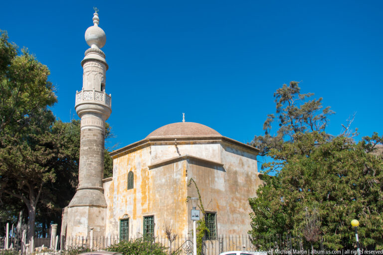 Small mosque with a red-brown dome and tall cylindrical stone minaret surrounded by mature trees.