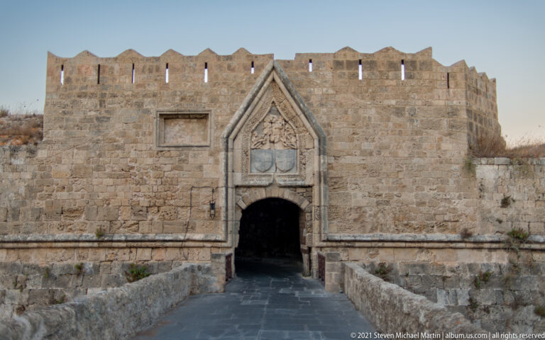 Fortified stone gateway with a pointed frame, carved heraldic panel, and narrow paved approach.