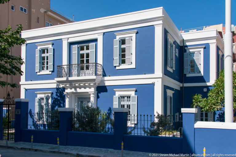 Two-story blue house with white trim, shutters, central iron balcony, and black street fence.