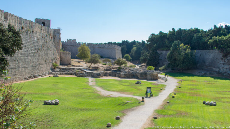 Grassy defensive moat between high stone walls, with walking paths, low ruins, and scattered stone balls.